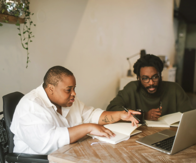 Two people, one in a wheelchair, sitting at a table with a laptop